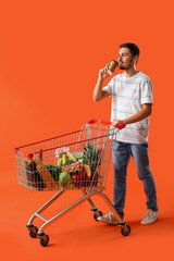 Young man with shopping cart and cup of coffee on orange background