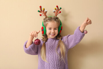 Happy little girl with Christmas baubles, reindeer antlers and earmuffs on beige background