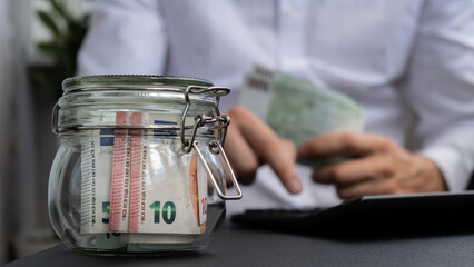 Man hands counting expenses banknotes of euro cash from glass jar in the piggy bank on calculator....