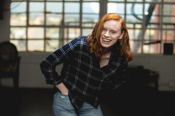 Young red haired woman in plaid shirt stands in an industrial space.