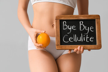 Beautiful young woman holding orange and board with text BYE BYE CELLULITE on grey background