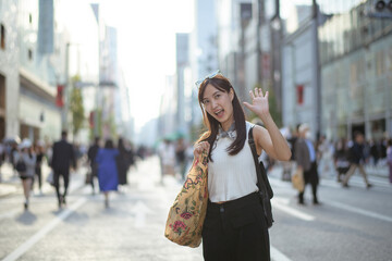 Asian woman in Tokyo's urban scene, standing at a road corner with a trendy tote bag, showcasing a...