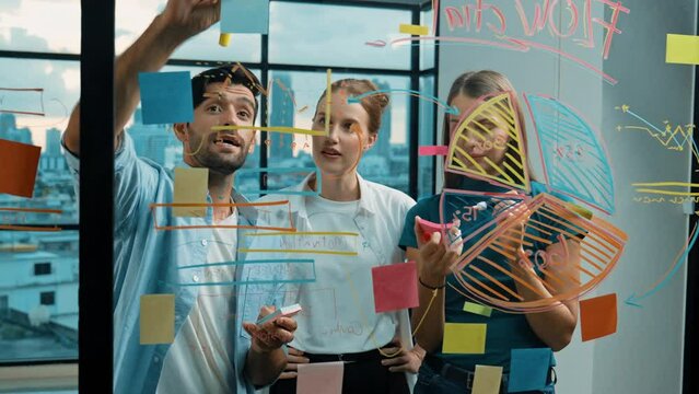 Group of professional business team working together, discussing and writing at sticky notes about marketing idea,plan, strategy at glass wall with graph and colorful sticky notes. Tracery