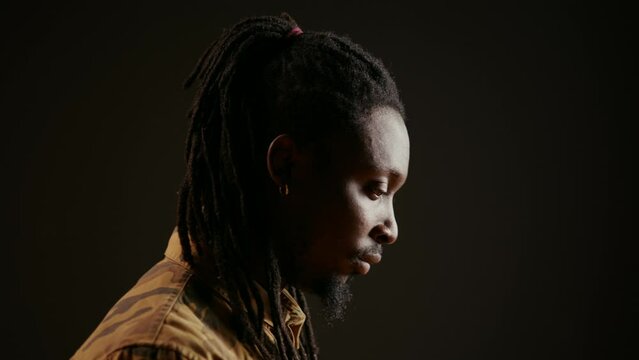 African american guy looking at camera with confidence, feeling happy and relaxed with dreads and cool clothing items. Model posing, standing over black background smiling. Handheld shot.