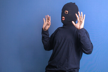 Thief wearing black mask raising hands up isolated over blue background