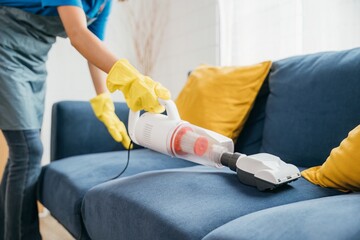 Asian housekeeper cheerfully vacuum machine cleaners a sofa focusing on hygiene and cleanliness in a living room. Her modern approach to cleaning furniture reflects dedication to her housework.