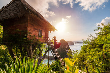 Young woman traveler enjoying and looking beautiful sunrise at the tree house in Nusa Penida island Bali, Indonesia - obrazy, fototapety, plakaty