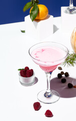 A delicate raspberry margarita cocktail, accented with fresh raspberries and pine cone decor on a geometric white stand