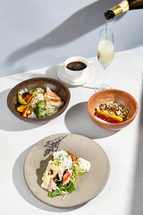 A modern breakfast arrangement with homemade yogurt, a chicken and avocado bowl, crab with a poached egg, served with coffee and sparkling wine being poured