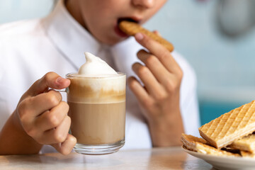 young girl is eating waffle and drinking cappuccino coffee with white foam. . Morning coffee and sweets in hands. - obrazy, fototapety, plakaty