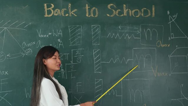 Back to school concept. Asian female teacher smiling with wooden stick pointing to blackboard at school in classroom, Happy beautiful young woman standing hold pointer to back board