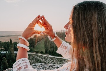 Obraz premium Happy woman in white boho dress making heart sign with hands on sunset in mountains. Romantic woman with long hair standing with her back on the sunset in nature in summer with open hands.