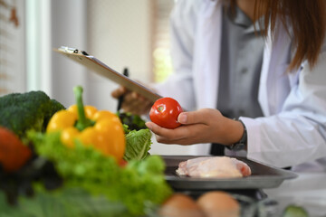 Professional nutritionist in white coat working at desk with vegetables. Healthcare and diet concept - obrazy, fototapety, plakaty