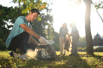 Smiling Asian male volunteer and his colleagues picking up plastic trash in public park - obrazy, fototapety, plakaty