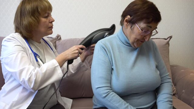 A home doctor massages the shoulders and back of an elderly woman using an electric massager. Ideal for content about senior healthcare, senior health care, and home health care programs.