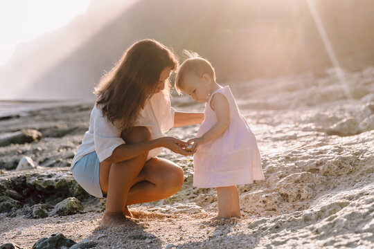 Cheerful mom teaches a child to walk on beach with sunset tones. Beautiful family on coastline