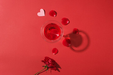 Red background featured a cocktail glass filled with red wine. A branch of rose and rose petals are decorated. Concept of Valentine’s Day for couples