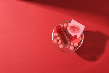 A red background with a glass full with pink chocolates, red paper heart and a lipstick unlabeled displayed. Top view and space for design. Valentine day greeting concept
