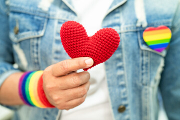 Asian lady wearing rainbow flag wristbands and hold red heart, symbol of LGBT pride month celebrate annual in June social of gay, lesbian, bisexual, transgender, human rights. - obrazy, fototapety, plakaty