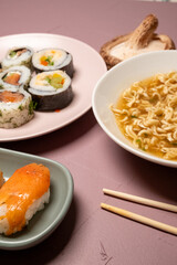 Chop sticks, mushrooms, noodle ramen, California rolls and Philadelphia rolls on a small plate on a pink background
