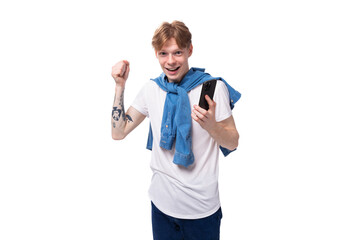 young fashionable caucasian guy with short red hair with a tattoo on his arms rejoices holding the phone