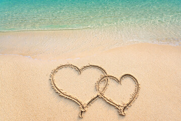 Two hearts handwritten on sandy beach with ocean wave on background. Two hearts drawn on sand with clear blue sea, copy space. Concept of luxury Honeymoon travel and greetings for valentine's day.