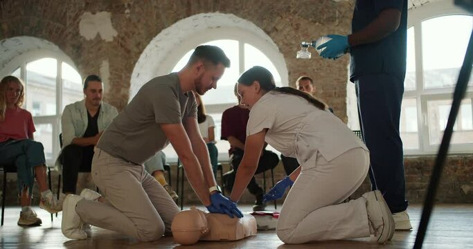 bearded brunette man in a gray T-shirt with a nurse girl in a white uniform demonstrates the rules of first aid with a mannequin in front of the public against the background of