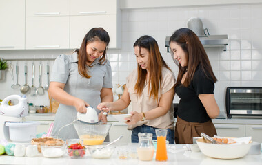 Asian happy professional female pastry bakery baker chef housewife with close friends standing smiling helping using eggs whisk beater machine mixing flour preparing dough in full decorated kitchen