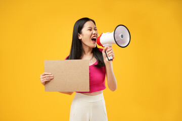 Portrait of a young Asian woman shouting angrily using a megaphone and holding paper. Isolated on a yellow background. Demonstration concept. 