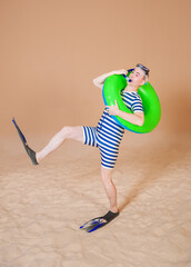 Funny man in vintage style striped swimsuit, inflatable ring, diving mask, snorkel and flippers walking on sand