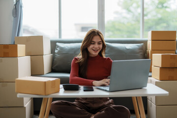 Smiling young business owner woman prepare parcel box and standing check online orders of product for deliver to customer on laptop computer. Shopping Online concept.