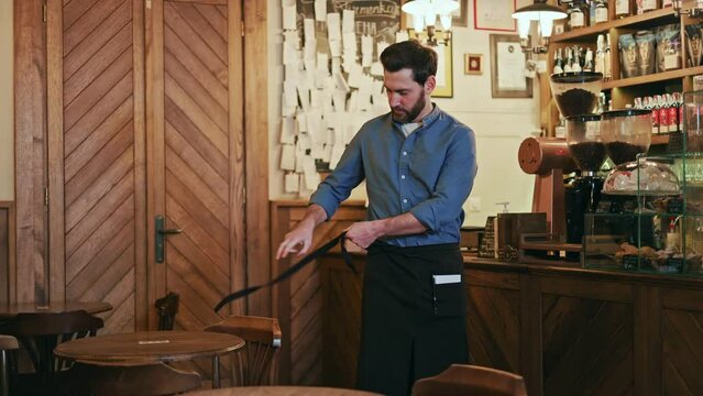 Portrait of handsome bearded waiter adeptly fastening black apron around waist and securing it with bow indoors. Caucasian food service worker preparing for workday in restaurant of retro style.