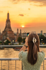 Naklejka premium tourist woman enjoys view to Wat Arun Temple in sunset, Traveler take photo to Temple of Dawn by smartphone from rooftop bar. Landmark and Travel destination in Bangkok, Thailand and Southeast Asia