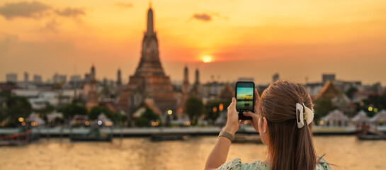 tourist woman enjoys view to Wat Arun Temple in sunset, Traveler take photo to Temple of Dawn by smartphone from rooftop bar. Landmark and Travel destination in Bangkok, Thailand and Southeast Asia - obrazy, fototapety, plakaty