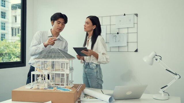 Beautiful young caucasian project manager brief asian engineer by using tablet while engineer measuring house model on meeting table with blueprint, architectural equipment scatter around. Immaculate