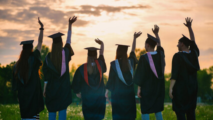 Silhouettes of graduates in black robes waving their arms against the evening sunset.