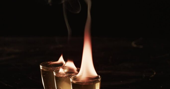 Two shot glasses with a clear liquid are ablaze with flames, set against a dark background