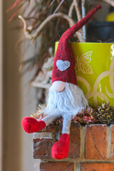 A Christmas gnome, bearded and wearing a long red hat, sits on the edge of a low brick wall. He's leaning against a green flowerpot.
