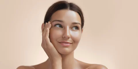 Skincare and Beauty. Woman with beautiful face touching healthy facial skin portrait. Young adult smiling Asian girl model with natural makeup touching glowing hydrated skin on beige background