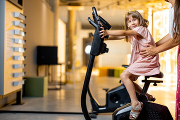 Little girl with mom training on bicycle simulator, visiting science museum. Concept of children's...