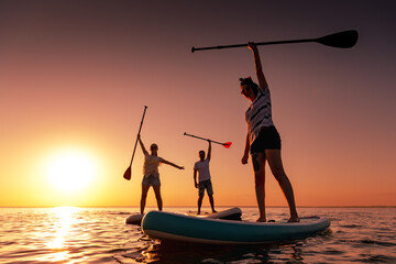 Three happy frieSilhouettes of three friends are standing in calm lake with sup boards in handsnds...