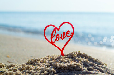Plastic stick in shape of red heart and word Love in sand on beach seashore on sunny summer day...