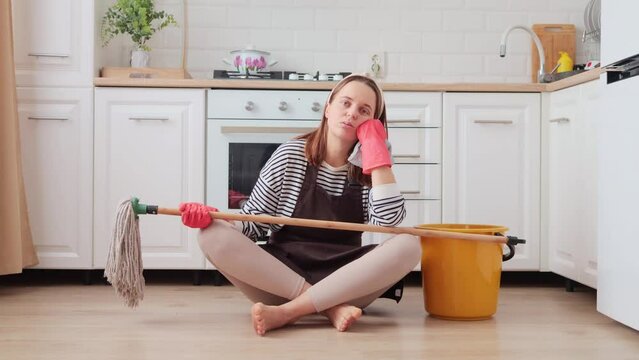 Sleepy yawning young adult Caucasian woman sitting near bucket with cleaning stuff at the domestic kitchen being tired and exhausted doing lots work about the house