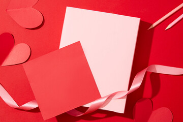 Paper cards, paper hearts, ribbons and candles are displayed on a red background. Ideal space to design wishes. Valentine's Day theme for advertising.