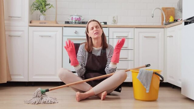 Frustrated sad helpless young woman sitting near bucket with cleaning stuff at domestic kitchen floor being tired and exhausted creaming with negative feelings