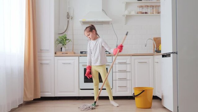 Home wet cleaning. Bored little girl in household rubber gloves washing floor with modern mop in kitchen child tidying up kitchen having unhappy expression