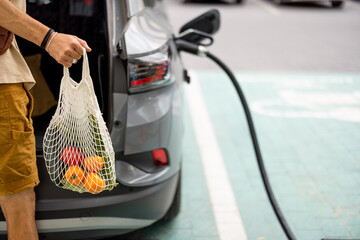 Man puts a bag full of fresh groceries in electric vehicle trunk, coming from the supermarket to...