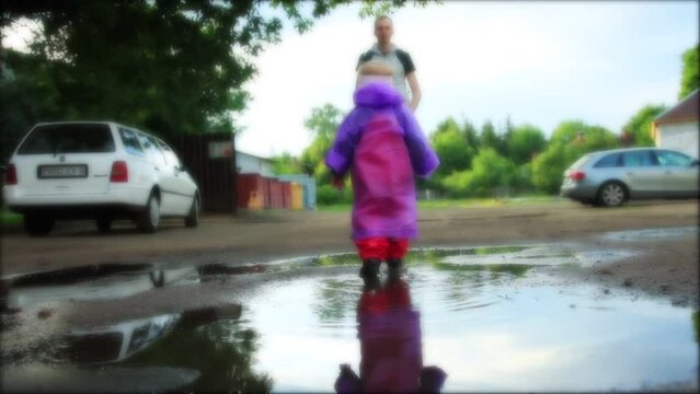 A little girl in a purple raincoat and rubber boots walks through the puddles after waterfall in the summer, background. Blurred, slow motion