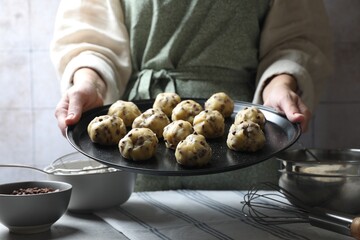 Woman holding tray with raw chocolate chip cookie balls above table indoors, closeup