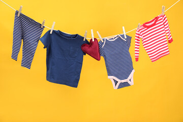 Different baby clothes and heart shaped cushion drying on laundry line against orange background